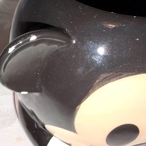 May include: A white ceramic mug with a black handle and the text "But First, Coffee" in a bold, black font. The mug is filled with a dark liquid, likely coffee, and sits on a light-colored surface.