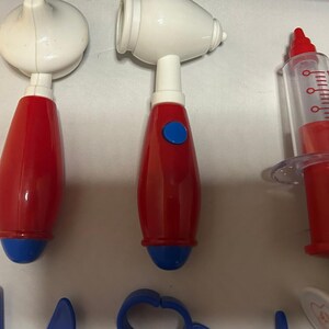 May include: A collection of toy medical tools, including a red and white stethoscope, an otoscope, and a syringe. The tools are primarily red and white with blue accents, designed for children's play.