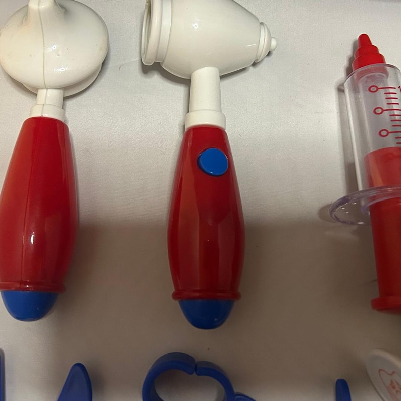 May include: A collection of toy medical tools, including a red and white stethoscope, an otoscope, and a syringe. The tools are primarily red and white with blue accents, designed for children's play.