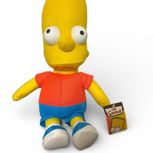 May include: A Bart Simpson soft toy with a yellow head, orange top, blue shorts, and white and blue trainers. The doll has large, white eyes and a small tag that reads "The Simpsons".