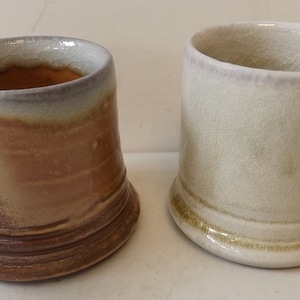 Puede incluir: Dos tazas de cerámica hechas a mano. Una es marrón con un degradado de colores, la otra es blanquecina. Ambas tienen asas curvas y una forma ligeramente irregular, lo que sugiere que están hechas a mano.