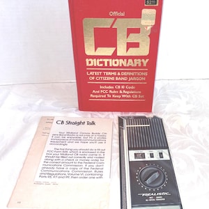 Könnte beinhalten: Ein rotes Buch "Official CB Dictionary" mit goldfarbenen Buchstaben, eine Broschüre "CB Straight Talk" und ein schwarz-silbernes "Realistic" CB-Funkgerät. Das Wörterbuch enthält den CB 10 Code und FCC-Regeln.