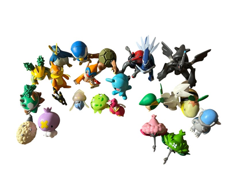 May include: A collection of various small plastic character figurines in diverse colors including blue, green, yellow, pink, and black. The figures feature distinct shapes, some with wings, spikes, or rounded bodies, arranged in a loose group.