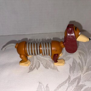 Puede incluir: Un juguete de perro salchicha con un cuerpo en forma de resorte. El perro es principalmente marr&oacute;n con una oreja roja, un hocico beige y patas amarillas. El cuerpo est&aacute; conectado por un resorte gris. Un juguete cl&aacute;sico para ni&ntilde;os.