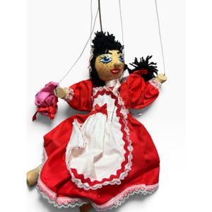 May include: A wooden marionette puppet with black yarn hair, blue eyes, and freckles, wearing a red dress with a white apron featuring lace trim and a small red bow. The puppet is suspended by thin strings and holds a small pink fabric accessory.