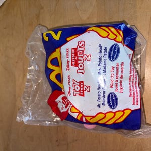 May include: A sealed plastic package featuring a Toy Story 2 McDonald's toy. The packaging is blue with yellow accents and the text "Toy Story 2" and "Jouets 2". The image of Mr. Potato Head is visible.