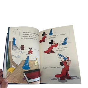 May include: Open book with illustrations of Mickey Mouse in a red robe and a blue sorcerer's hat. Text on the page reads, "Now I can be a sorcerer," said Mickey. The book is open to a double-page spread with colorful drawings.