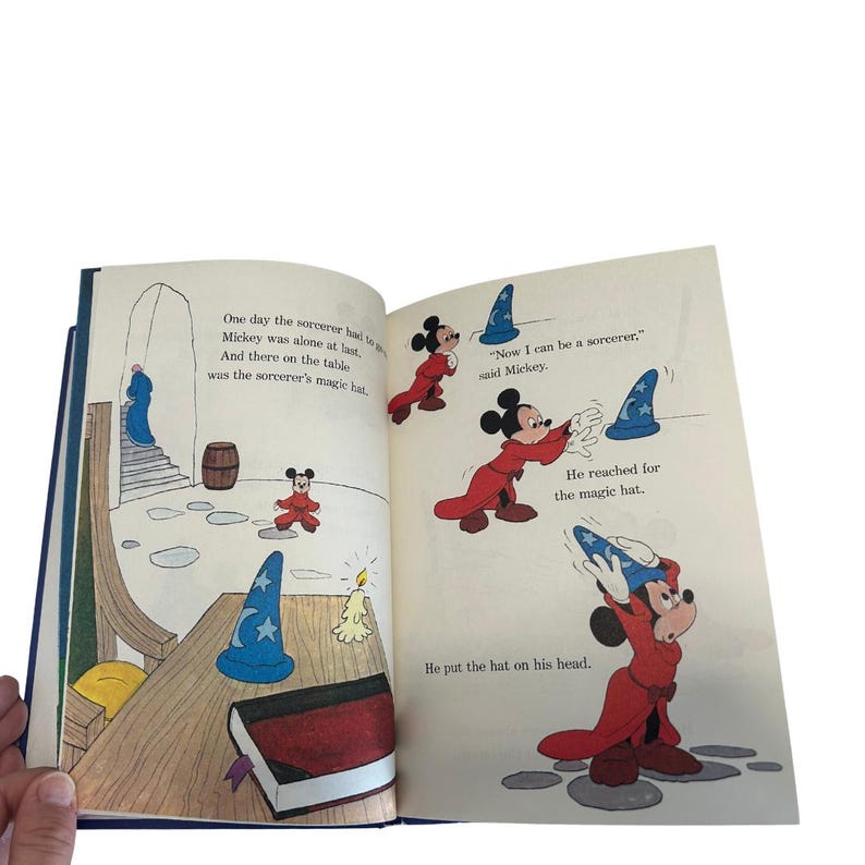 May include: Open book with illustrations of Mickey Mouse in a red robe and a blue sorcerer's hat. Text on the page reads, "Now I can be a sorcerer," said Mickey. The book is open to a double-page spread with colorful drawings.