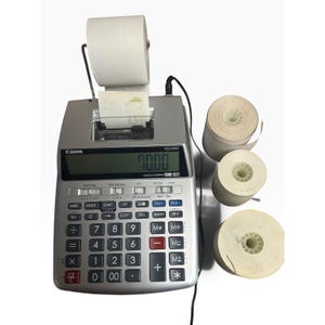 Puede incluir: Una calculadora impresora Canon P23-DHV plateada con pantalla digital que muestra 70.00, soporte para rollo de papel y cable de alimentación negro. Tres rollos de papel blanco adicionales están situados a la derecha de la calculadora.