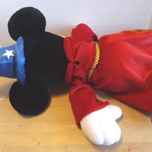 May include: A plush toy of a black-eared character wearing a blue wizard hat with white stars and a red robe. The robe is tied with a gold rope. The character has white gloves and brown feet.