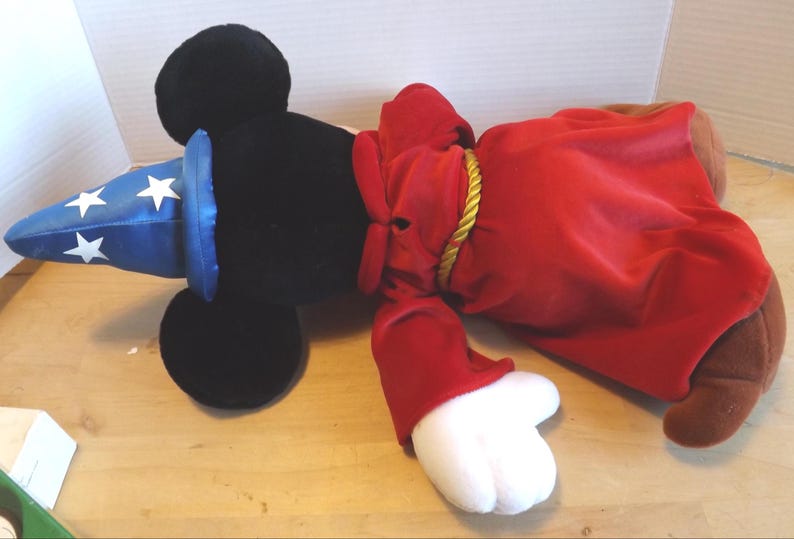 May include: A plush toy of a black-eared character wearing a blue wizard hat with white stars and a red robe. The robe is tied with a gold rope. The character has white gloves and brown feet.