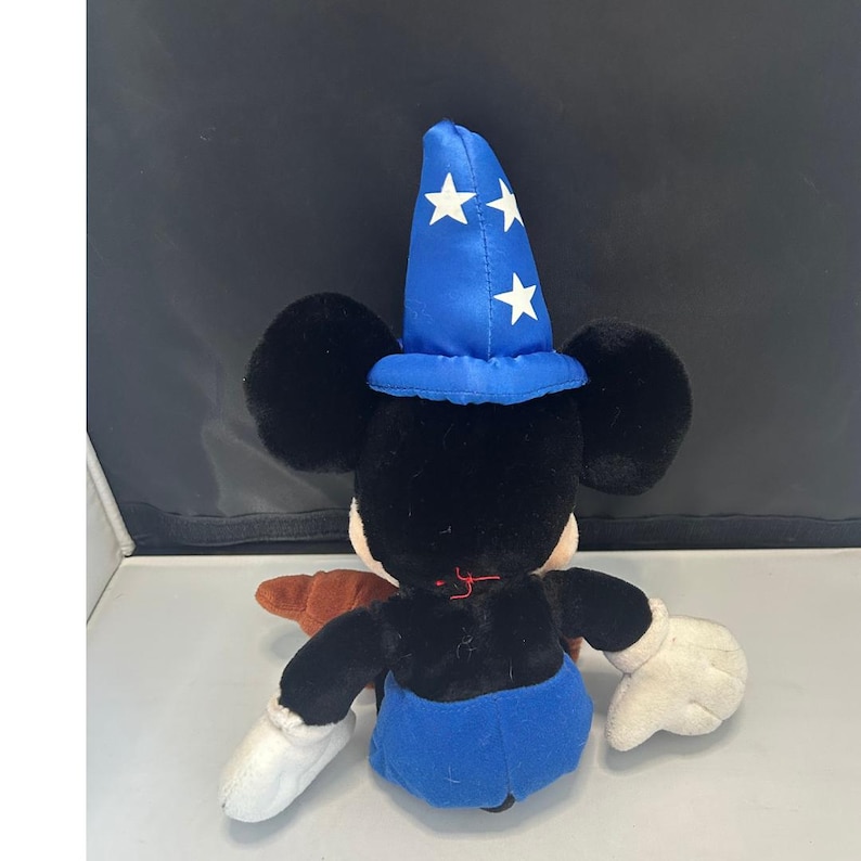 May include: A plush Mickey Mouse toy, viewed from the back, wearing a blue wizard hat with white stars. The toy has black ears, a black body, blue shorts, and white gloves. A brown object is visible on the left side.