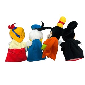 以下が含まれることがあります： 白雪姫、ドナルドダック、グーフィー、ミッキーマウスの4つのディズニーキャラクターの手人形。人形は柔らかい布でできており、鮮やかな色をしています。白雪姫は黄色の帽子と赤いマントを着ています。ドナルドダックは青いセーラーハットをかぶっています。