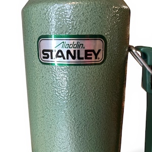 May include: A green Stanley flask with a textured finish. The flask features a handle and a silver and green label displaying "Aladdin STANLEY". Designed to maintain liquid temperature, it's a classic item.