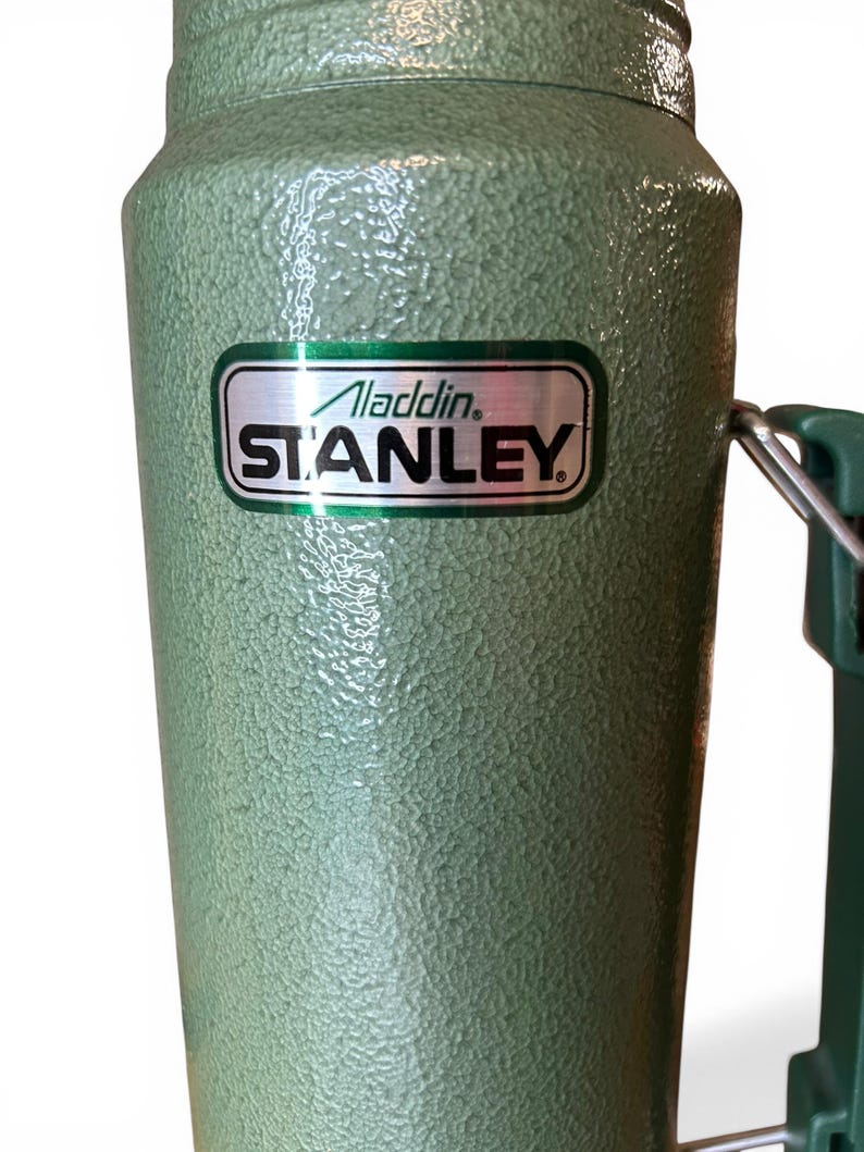 May include: A green Stanley flask with a textured finish. The flask features a handle and a silver and green label displaying "Aladdin STANLEY". Designed to maintain liquid temperature, it's a classic item.
