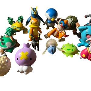 May include: A collection of various small plastic character figurines featuring diverse shapes and colors, including a round purple creature with a yellow cross, a blue penguin, a green turtle-like figure, and several other stylized animal-like beings.