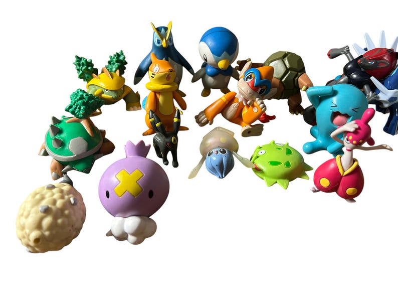 May include: A collection of various small plastic character figurines featuring diverse shapes and colors, including a round purple creature with a yellow cross, a blue penguin, a green turtle-like figure, and several other stylized animal-like beings.