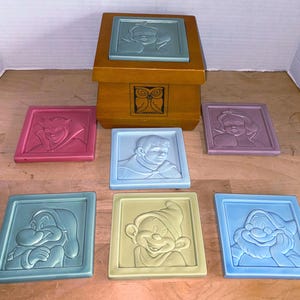 Op de afbeelding: Een set van zeven vierkante onderzetters met reliëfbeelden van Disney-personages. De onderzetters zijn in verschillende kleuren, waaronder roze, blauw en geel. Ze zijn gerangschikt rond een houten kist met een uilontwerp.