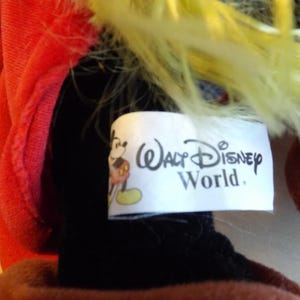 May include: Close-up of a plush toy with a red outfit and yellow hair. A white tag displays the text "Walt Disney World" along with a Mickey Mouse graphic. The toy's black and brown elements are visible.