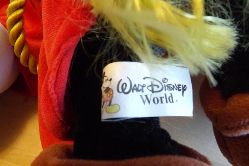 May include: Close-up of a plush toy with a red outfit and yellow hair. A white tag displays the text "Walt Disney World" along with a Mickey Mouse graphic. The toy's black and brown elements are visible.