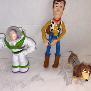 Puede incluir: Colecci&oacute;n de figuras de personajes de Toy Story, incluyendo a Buzz Lightyear con un traje espacial blanco y verde, Woody el vaquero con camisa amarilla y pantalones azules, y Slinky Dog, un perro salchicha marr&oacute;n con cuerpo de resorte.