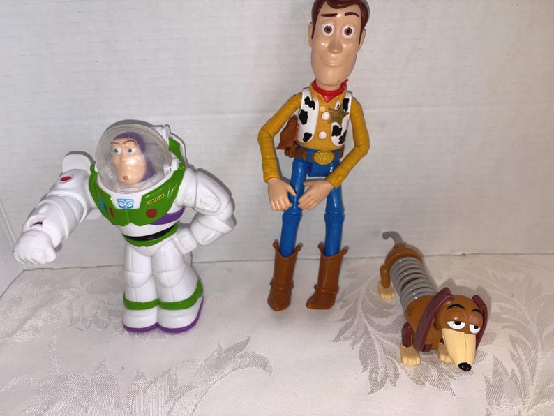 Puede incluir: Colecci&oacute;n de figuras de personajes de Toy Story, incluyendo a Buzz Lightyear con un traje espacial blanco y verde, Woody el vaquero con camisa amarilla y pantalones azules, y Slinky Dog, un perro salchicha marr&oacute;n con cuerpo de resorte.