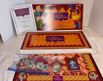 May include: A Disney's Hunchback of Notre Dame Town Square Game. The board game includes a game board, instructions, game pieces, and cards. The box is red and orange with character illustrations. The game pieces are in a clear plastic bag.
