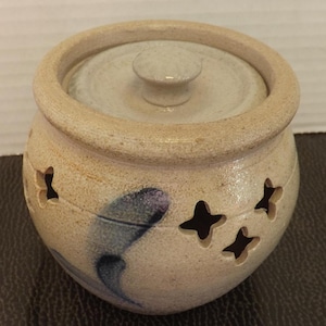 Puede incluir: Un pequeño tarro de cerámica beige con tapa y pomo. El tarro tiene recortes en forma de estrella y un diseño pintado en azul y morado. La tapa es de un tono beige más claro. El tarro es probablemente hecho a mano.