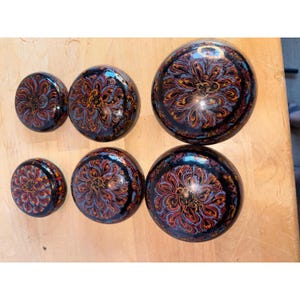 May include: A set of six round, black lacquer containers of varying sizes, arranged in two rows. Each container features a glossy finish and a detailed, hand-painted floral pattern in shades of red, blue, and gold, radiating from the center.