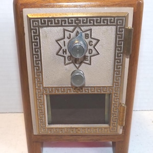 May include: A wooden mailbox with a light-colored metal door. The door features a starburst design with letters and a dial. The frame is trimmed with a gold-colored Greek key pattern. The mailbox has a small window and sits on four wooden legs.
