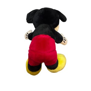 Puede incluir: Un peluche de Mickey Mouse, visto desde atr&aacute;s, presenta una cabeza y orejas negras, pantalones cortos rojos, zapatos amarillos y guantes blancos. El peluche est&aacute; dise&ntilde;ado con materiales suaves y afelpados.
