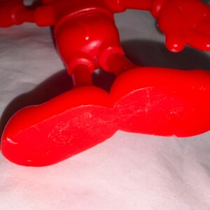 K&ouml;nnte beinhalten: Nahaufnahme einer hellroten Kunststoff-Spielfigur. Die gro&szlig;en, abgerundeten F&uuml;&szlig;e der Figur befinden sich im Vordergrund, der Rest des K&ouml;rpers und die Arme sind dar&uuml;ber zu sehen. Das Spielzeug befindet sich auf einer wei&szlig;en Oberfl&auml;che.