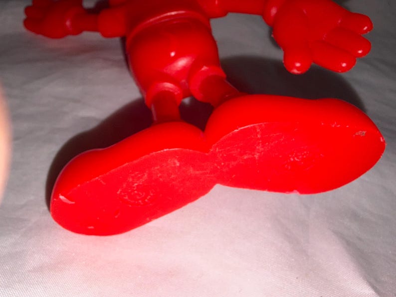 K&ouml;nnte beinhalten: Nahaufnahme einer hellroten Kunststoff-Spielfigur. Die gro&szlig;en, abgerundeten F&uuml;&szlig;e der Figur befinden sich im Vordergrund, der Rest des K&ouml;rpers und die Arme sind dar&uuml;ber zu sehen. Das Spielzeug befindet sich auf einer wei&szlig;en Oberfl&auml;che.