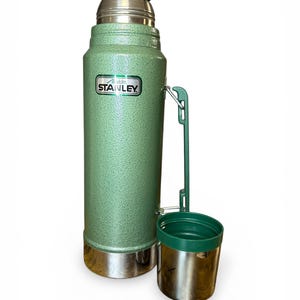 May include: A green Stanley flask with a silver base and a green lid. The flask has a matching green cup attached. The Stanley logo is visible on the front. Designed to keep liquids hot or cold. Measures approximately 30 cm in height.