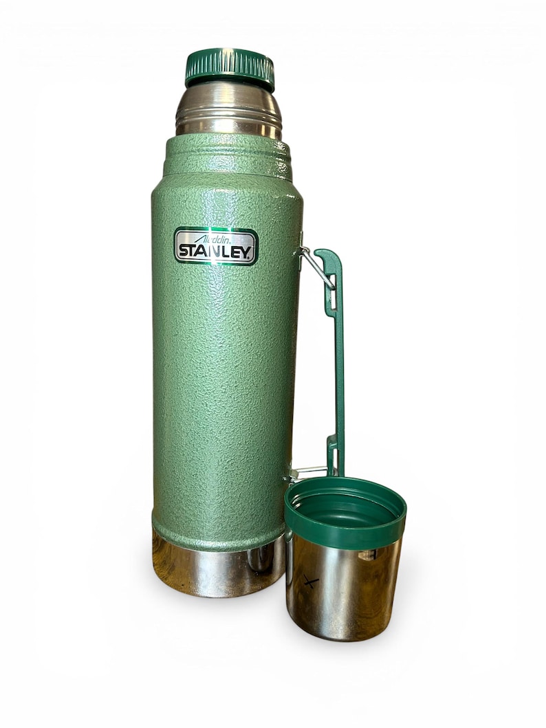 May include: A green Stanley flask with a silver base and a green lid. The flask has a matching green cup attached. The Stanley logo is visible on the front. Designed to keep liquids hot or cold. Measures approximately 30 cm in height.