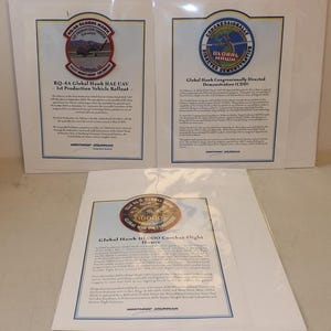 May include: Three framed documents with text and logos related to the Global Hawk UAV. The top left document features a patch with an aircraft and the text "RQ-4A Global Hawk HAE UAV". The top right document has a patch with the text "Global Hawk Congressionally Directed Demonstration". The bottom document has a patch with the text "Global Hawk 10,000 Combat Flight Hours".