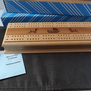 Op de afbeelding: Een lichtbruin houten cribbagebord met een contrasterende houten inleg, herten silhouetten en de tekst "Friendship Hardwoods". Inclusief een blauw-wit gestreepte doos, een zwarte kunstleren mat en een kaart met "Handmade In The USA".