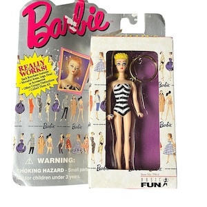 May include: A miniature Barbie doll keychain in original packaging. The doll has blonde hair and wears a black and white striped swimsuit. The packaging features the Barbie logo, illustrations of various outfits, and text including "REALLY WORKS!" and "WARNING: CHOKING HAZARD."