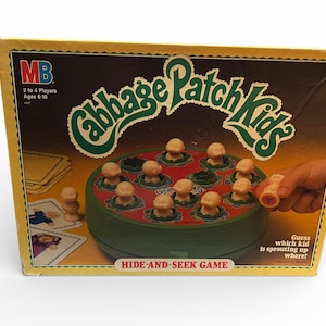 Könnte beinhalten: Vintage Cabbage Patch Kids Versteckspiel. Die Box ist gelb und braun mit dem Spieltitel in Grün und Weiß. Das Spiel zeigt kleine Plastikpuppen und ein kreisförmiges Spielbrett. Der Text auf der Box lautet "2 bis 4 Spieler, Alter 6-10".