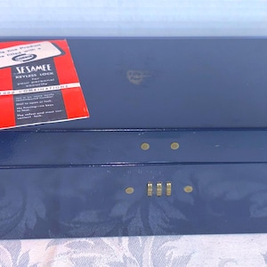 May include: A navy blue rectangular box with a hinged lid. The box has a combination lock and brass-colored hardware. A red tag with white text is attached to the box. The text on the tag reads "SESAMEE".