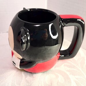 May include: A ceramic mug featuring a cartoon character design. The mug is primarily black with red and white accents, including a red handle. The mug has a glossy finish and a rounded shape.