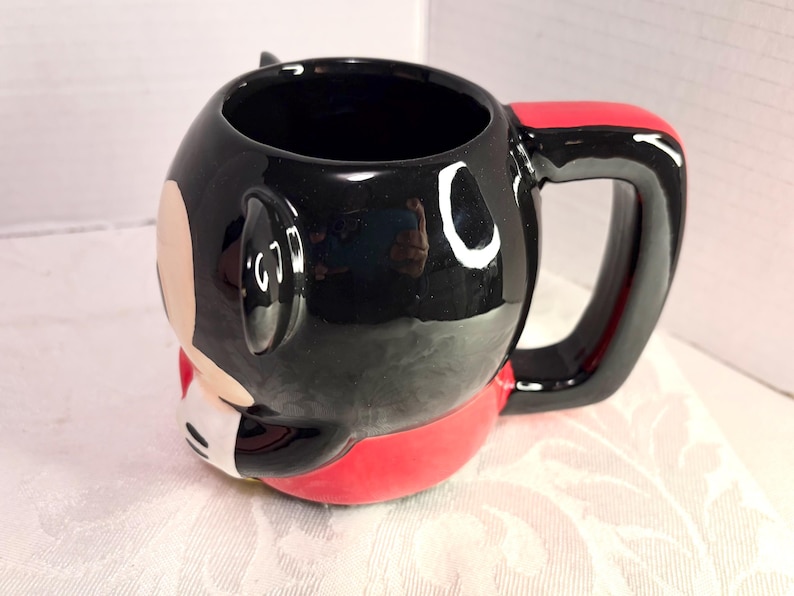May include: A ceramic mug featuring a cartoon character design. The mug is primarily black with red and white accents, including a red handle. The mug has a glossy finish and a rounded shape.