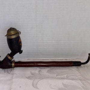May include: A vintage tobacco pipe with a dark brown, curved stem and a black bowl with a hinged, brass-colored lid. The bowl has a decorative design. The pipe is resting on a white surface.