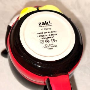 May include: A black, red, and yellow ceramic mug with a handle. The bottom of the mug has the text "zak!", "Disney", "HAND WASH ONLY", and "13+". The mug is from 2019 and made in China.