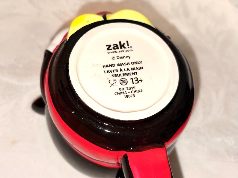May include: A black, red, and yellow ceramic mug with a handle. The bottom of the mug has the text "zak!", "Disney", "HAND WASH ONLY", and "13+". The mug is from 2019 and made in China.