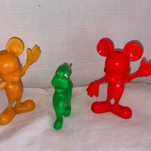 Könnte beinhalten: Drei Plastikfiguren: eine orangefarbene Maus mit erhobenen Armen, ein grünes dinosaurierartiges Wesen und eine rote Maus mit erhobenen Armen. Die Figuren stehen auf einer weißen Oberfläche.