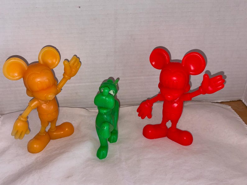 K&ouml;nnte beinhalten: Drei Plastikfiguren: eine orangefarbene Maus mit erhobenen Armen, ein gr&uuml;nes dinosaurierartiges Wesen und eine rote Maus mit erhobenen Armen. Die Figuren stehen auf einer wei&szlig;en Oberfl&auml;che.