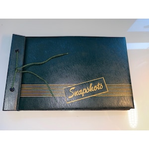 May include: A rectangular dark green photo album with a textured cover. The front features gold horizontal lines and the word Snapshots in a gold script font. A dark green cord is threaded through two eyelets on the left side of the album.