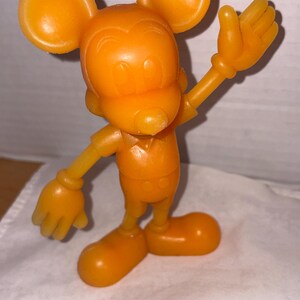 K&ouml;nnte beinhalten: Orangefarbene Mickey Mouse-Figur mit gro&szlig;en, runden Ohren, einem l&auml;chelnden Gesicht und ausgestreckten Armen. Das Vintage-Spielzeug steht auf einer wei&szlig;en Oberfl&auml;che und besteht aus einem festen, glatten Material. Die Figur ist eine klassische Zeichentrickfigur.