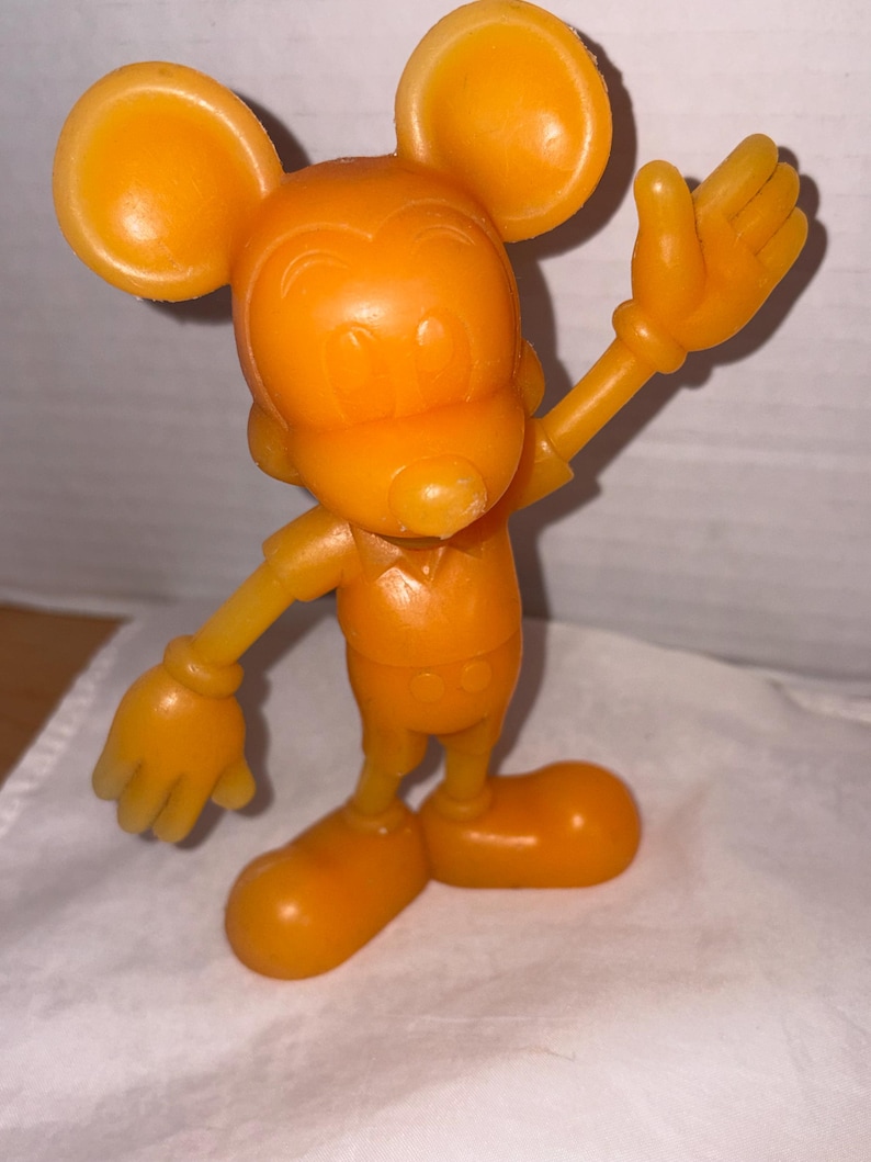 K&ouml;nnte beinhalten: Orangefarbene Mickey Mouse-Figur mit gro&szlig;en, runden Ohren, einem l&auml;chelnden Gesicht und ausgestreckten Armen. Das Vintage-Spielzeug steht auf einer wei&szlig;en Oberfl&auml;che und besteht aus einem festen, glatten Material. Die Figur ist eine klassische Zeichentrickfigur.